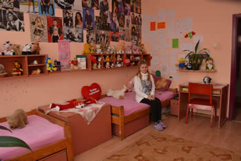 The child is sitting on the bed in the room of Yefimija's home. The walls are orange and decorated with posters in the area above the bed.